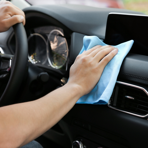A hand holds a folded blue microfiber cloth, wiping the top of a black car dashboard and air vent. The driver's steering wheel is visible on the left. Product used is Ultra Shine Protectant.