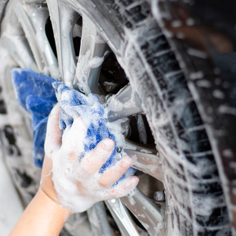 A hand in a blue cloth is shown scrubbing a dirty car wheel covered in thick, white foam. The hand is cleaning an alloy rim and the side of the black tire. Product used is Wheel & Tire Cleaner Concentrate.